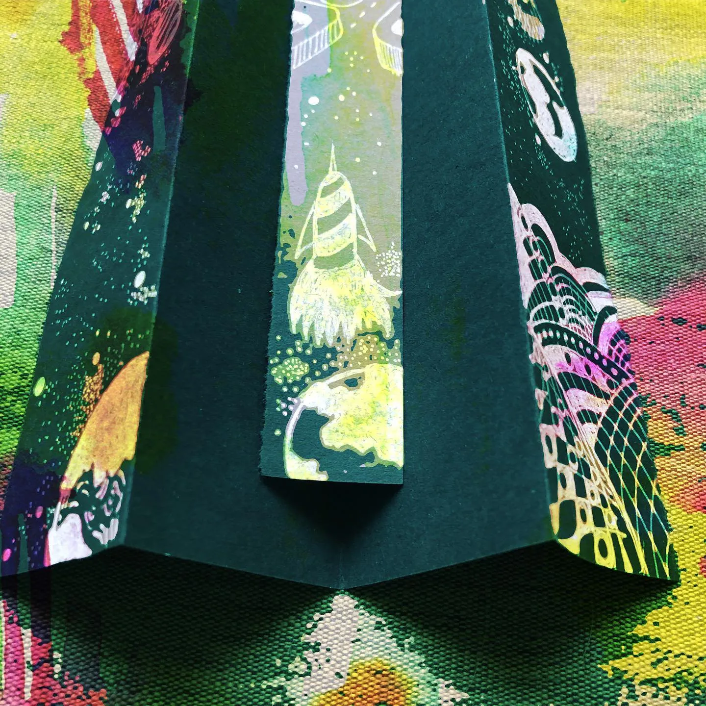 A dark green card folded into pleats like a tent, holding the white ink rocket bookmark at its centre, standing on a canvas splashed with bright yellow, pink and green paint.